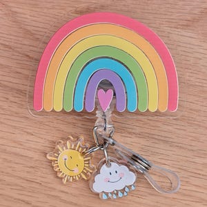 Peut inclure: Un enrouleur de badge arc-en-ciel color&eacute; avec un c&oelig;ur, un soleil et un nuage. L'arc-en-ciel pr&eacute;sente des arches roses, oranges, jaunes, vertes, bleues et violettes. Les breloques soleil et nuage ont des visages souriants. Le reel a une sangle en plastique transparent et un clip en m&eacute;tal.