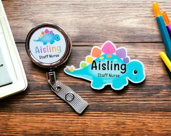 May include: A blue and rainbow dinosaur-shaped name tag with the text "Aisling Staff Nurse". The name tag is attached to a silver retractable badge holder with a gray strap.