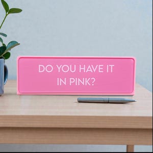 Pu&ograve; includere: Un cartello rettangolare rosa acceso con il testo bianco "DO YOU HAVE IT IN PINK?". Una penna d'argento &egrave; appoggiata su un tavolo di legno davanti al cartello.