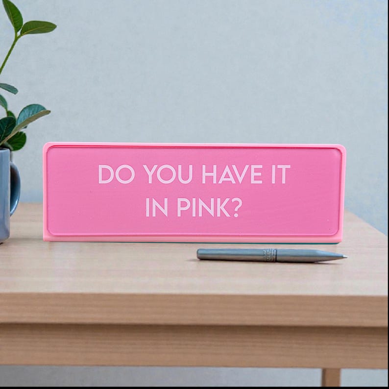 Pu&ograve; includere: Un cartello rettangolare rosa acceso con il testo bianco "DO YOU HAVE IT IN PINK?". Una penna d'argento &egrave; appoggiata su un tavolo di legno davanti al cartello.