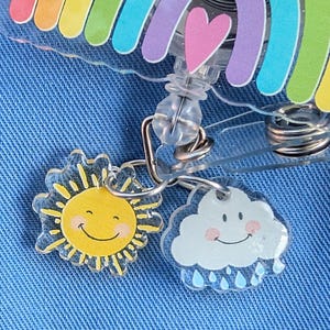 Peut inclure: Un enrouleur de badge en acrylique transparent avec un motif arc-en-ciel et un c&oelig;ur rose. Des breloques repr&eacute;sentant un soleil souriant et un nuage souriant avec des gouttes de pluie sont attach&eacute;es. Sur fond de denim bleu.