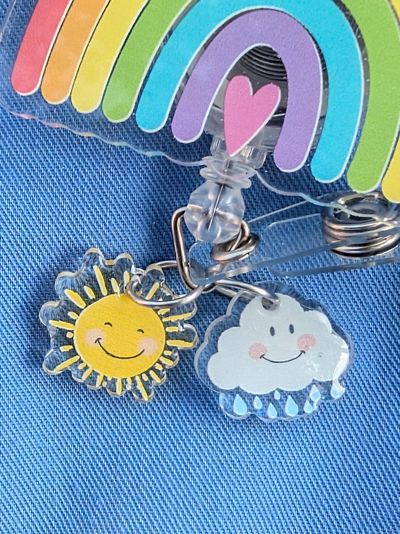 Peut inclure: Un enrouleur de badge en acrylique transparent avec un motif arc-en-ciel et un c&oelig;ur rose. Des breloques repr&eacute;sentant un soleil souriant et un nuage souriant avec des gouttes de pluie sont attach&eacute;es. Sur fond de denim bleu.