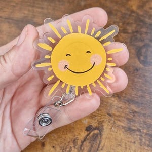 Op de afbeelding: Een gele en transparante badge reel met een lachend zonontwerp. De zon heeft gele stralen, een gele gezicht, roze wangen en een zwarte glimlach. De badge reel heeft een transparant intrekbaar plastic koord en een metalen clip.