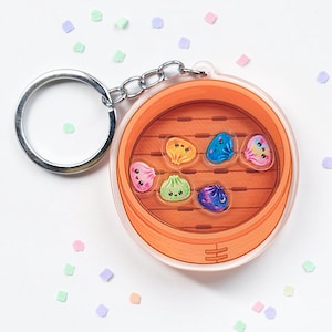 May include: A circular acrylic keyring featuring a printed illustration of a bamboo steamer basket containing six small, colourful, cartoon-style dumplings with smiling faces in shades of pink, orange, green, blue, and purple, attached to a metal ring.