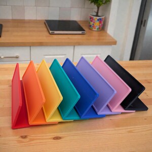 Pu&ograve; includere: Una fila di supporti per tablet colorati e angolati in un arcobaleno di tonalit&agrave;: rosso, arancione, giallo, verde, blu, viola, rosa e nero. I supporti sono disposti su una superficie di legno, mostrando un vibrante spettro di colori.