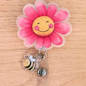 May include: A daisy-shaped badge reel with a smiling face and pink petals. A small bee-shaped charm is attached to the reel. The badge reel has a clear plastic retractable cord and a metal clip.