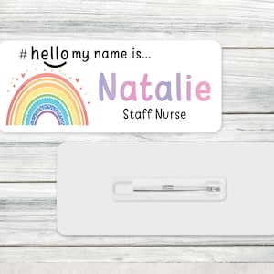 May include: A white name tag with a rainbow and the text "#hello my name is... Natalie Staff Nurse". The tag has a pin on the back.