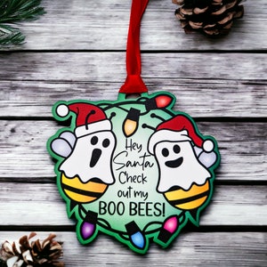 May include: A green Christmas bauble featuring two ghosts wearing Santa hats and bee wings. The bauble has the text "Hey Santa Check out my BOO BEES!" and is decorated with colourful lights.