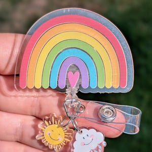 Peut inclure: Un porte-badge en acrylique transparent avec un motif arc-en-ciel color&eacute; et un c&oelig;ur rose. Des breloques repr&eacute;sentant un soleil souriant et un nuage avec des gouttes de pluie sont attach&eacute;es. Le porte-badge a une sangle en plastique transparent et un clip en m&eacute;tal.
