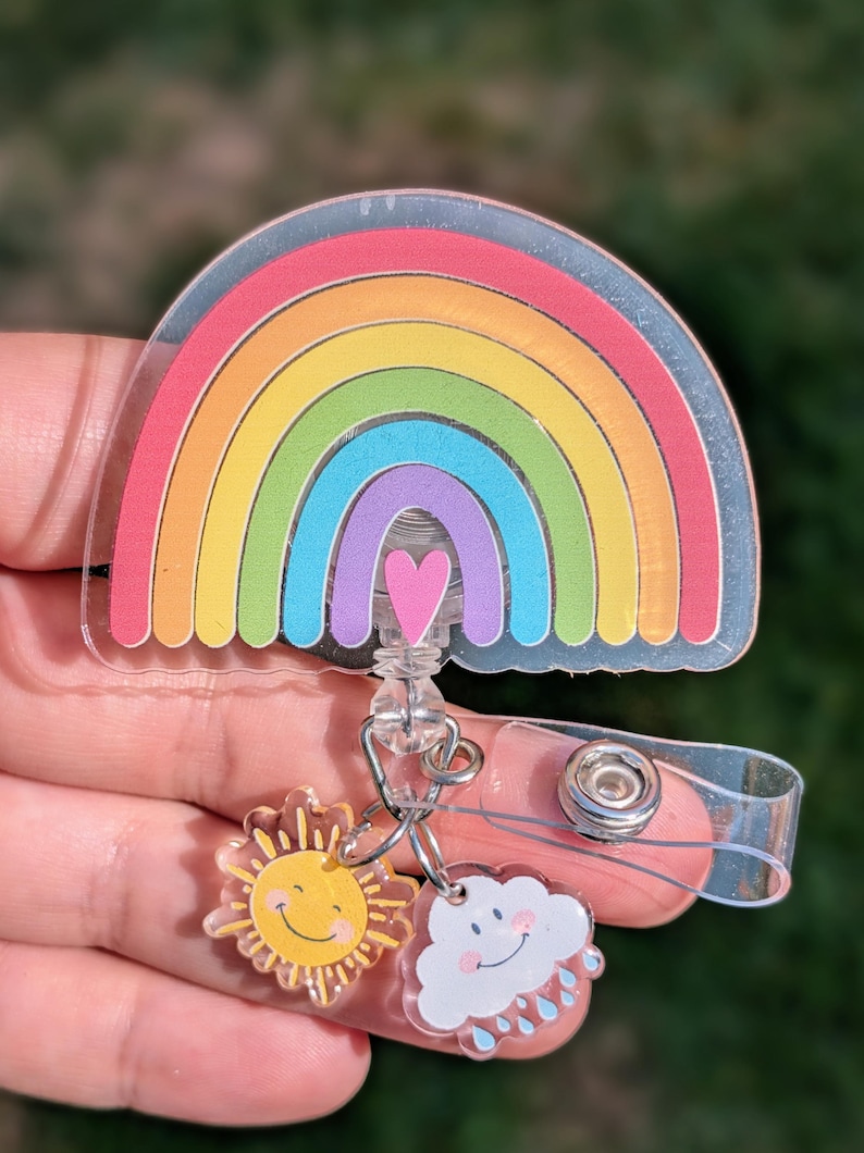 Peut inclure: Un porte-badge en acrylique transparent avec un motif arc-en-ciel color&eacute; et un c&oelig;ur rose. Des breloques repr&eacute;sentant un soleil souriant et un nuage avec des gouttes de pluie sont attach&eacute;es. Le porte-badge a une sangle en plastique transparent et un clip en m&eacute;tal.