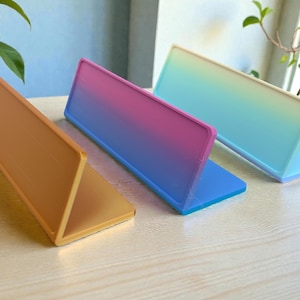Pu&ograve; includere: Tre espositori rettangolari colorati. Uno &egrave; arancione, un altro ha una sfumatura da rosa a blu, e il terzo ha una sfumatura da verde a bianco. Ogni espositore &egrave; piegato a forma triangolare, appoggiato su una superficie di legno chiaro.