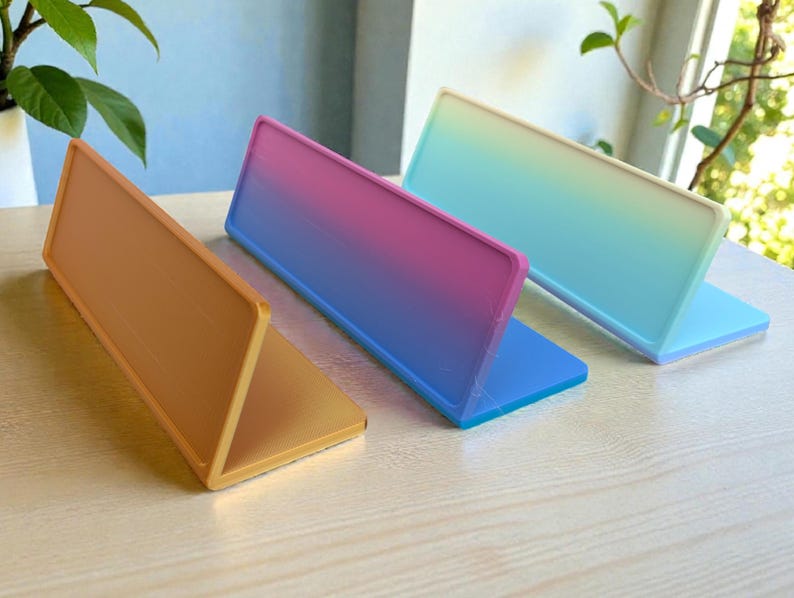 Pu&ograve; includere: Tre espositori rettangolari colorati. Uno &egrave; arancione, un altro ha una sfumatura da rosa a blu, e il terzo ha una sfumatura da verde a bianco. Ogni espositore &egrave; piegato a forma triangolare, appoggiato su una superficie di legno chiaro.