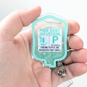 以下が含まれることがあります： グリッターで満たされた、ターコイズ色の縁取りがされた透明なIVバッグ型のバッジリール。「Pixie Dust Infusion」と「Contains Playful and Mischievous Fairy Energy」と書かれています。