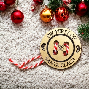 May include: A wooden ornament shaped like a button with the text "PROPERTY OF SANTA CLAUS" around the edge. The button has a gold centre with two red and white candy canes inside.