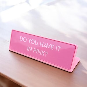 Pu&ograve; includere: Un cartello da scrivania rosa acceso con il testo bianco "DO YOU HAVE IT IN PINK?". Il cartello rettangolare ha un bordo leggermente rialzato ed &egrave; inclinato, appoggiato su una superficie di legno.