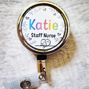 May include: A silver retractable badge holder with a white round insert featuring a colourful name "Katie" and the text "Staff Nurse" surrounded by a floral design with pink hearts.