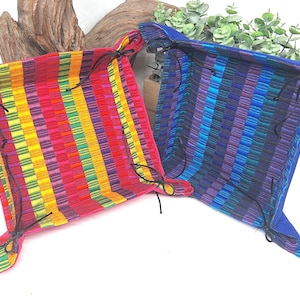 May include: Two square fabric trays with colorful striped patterns. One tray is red with yellow, green, and purple stripes. The other is blue with purple and teal stripes. Both have black ties at the corners.