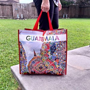 May include: A red tote bag with the word "GUATEMALA" in colorful letters. The bag features a vibrant, patterned design with images of buildings and cultural motifs. The bag has red handles and is held by a person.