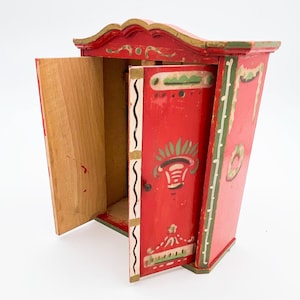 Könnte beinhalten: Ein roter Holzschrank mit einem bemalten Blumendesign und goldenen Akzenten. Der Schrank hat zwei Türen, von denen eine geöffnet ist und den leeren Innenraum zeigt.