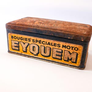 Può includere: Una scatola di metallo arrugginita con un'etichetta gialla e nera che recita "BOUGUIES SPECIALES MOTO EYQUEM".