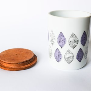 Puede incluir: Un tarro de cerámica blanca con una tapa de madera. El tarro tiene un patrón de hojas moradas y negras.