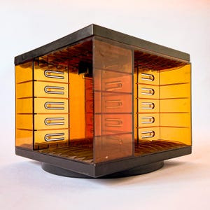 May include: A vintage, rotating CD storage cube. The cube is black with amber-coloured, transparent plastic panels. The interior features multiple slots for CD storage. The cube sits on a black, circular base.