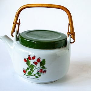 Könnte beinhalten: Weiße Teekanne aus Keramik mit grünem Deckel und Bambusgriff. Die Teekanne hat ein florales Design mit roten Beeren und grünen Blättern. Der Text "Rosa canina" ist auf der Unterseite der Teekanne aufgedruckt.