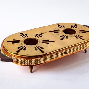 Pode incluir: Um aquecedor de alimentos de latão vintage com formato oval. Possui duas aberturas circulares na parte superior, recortes decorativos e alças de madeira. O aquecedor tem um padrão semelhante a uma malha nas laterais e repousa sobre pequenas pernas.