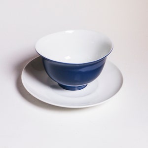 Könnte beinhalten: Eine weiße Untertasse mit einer blauen Teetasse darauf. Die Teetasse ist im traditionellen chinesischen Stil mit einer abgerundeten Form und einem breiten Rand.