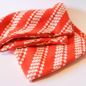 May include: Two folded orange and white woven fabric items with a zip. The fabric has a diagonal striped pattern with a checkered design. The items are likely a clutch or small bag.