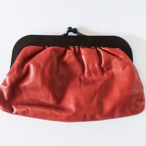 Puede incluir: Un clutch de cuero rojo con marco y cierre negros. El clutch tiene un diseño fruncido y una estética vintage. El bolso es de tamaño mediano y es adecuado para llevar lo esencial. El color es un rojo intenso y cálido.
