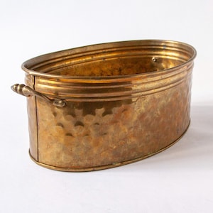 Puede incluir: Jardinera ovalada de latón con textura martillada y dos asas pequeñas. La jardinera tiene un tono dorado y un borde decorativo. Un recipiente de estilo vintage para plantas u otros objetos decorativos.