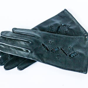Könnte beinhalten: Ein Paar schwarze Lederhandschuhe mit dekorativen Ausschnitten. Die Handschuhe sind in der Hälfte gefaltet, wobei die Fingerspitzen eines Handschuhs sichtbar sind.