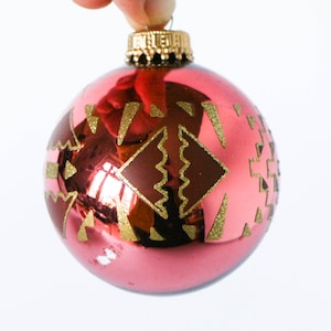 Könnte beinhalten: Ein roter gläserner Weihnachtsbaumschmuck mit einem goldenen geometrischen Muster. Der Schmuck ist rund und hat eine glänzende Oberfläche.