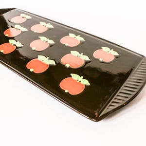 Könnte beinhalten: Ein braunes Keramik-Serviertablett mit erhöhtem Rand und einem sich wiederholenden Muster aus roten Äpfeln mit grünen Blättern. Das Tablett hat eine glänzende Oberfläche.