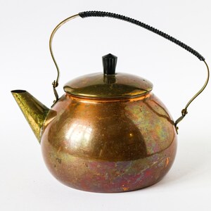 Könnte beinhalten: Eine Kupfer-Teekanne mit einem Messingdeckel und einem schwarzen Griff. Die Teekanne hat einen runden Körper und einen Ausguss an der Seite.