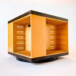 May include: A vintage, rotating storage unit in shades of tan and brown. The unit has a square shape with open sides and multiple slots for organizing items. The top and base are dark brown, and the unit sits on a black base.
