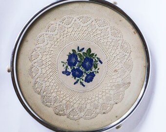 Op de afbeelding: Een ronde serveerschaal met een zilveren rand, een wit kantmotief en een blauw bloemenmotief in het midden.