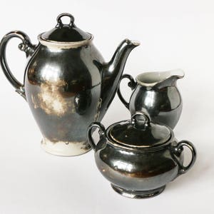 Könnte beinhalten: Ein silbernes Teeservice mit Teekanne, Milchkännchen und Zuckerdose. Das Set ist aus Porzellan und hat eine glänzende, metallische Oberfläche.