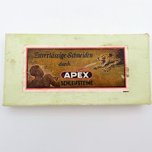 Könnte beinhalten: Eine Vintage-Schachtel mit Apex Schleifsteinen. Der Hintergrund ist grün und die Etikette zeigt einen Mann mit einem Speer und einen Leoparden. Auf der Etikette steht "Zuverlässige Schneiden durch APEX Schleifsteine".
