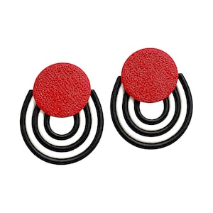 May include: A pair of earrings featuring a textured red circular top piece attached to a series of three concentric black and white semi-circular arches. The design consists of a solid red disc positioned above nested curved lines in black and white.