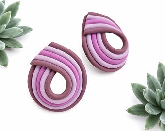 May include: A pair of teardrop-shaped earrings constructed from layered, curved tubes in varying shades of purple, ranging from light lavender to deep plum. The design features a continuous, looped structure with a smooth, matte finish.