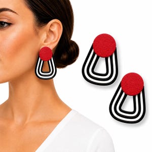 Puede incluir: Un par de pendientes con una pieza superior circular roja texturizada unida a un marco geométrico colgante. El marco consta de dos bucles en forma de U anidados en colores negro y blanco, creando un diseño lineal y superpuesto.