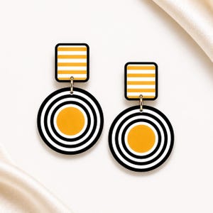 May include: A pair of geometric earrings with a modern design. Each earring features a rectangular top with horizontal yellow and white stripes, connected to a circular pendant with concentric black and white stripes and a yellow centre. The earrings have a black border.