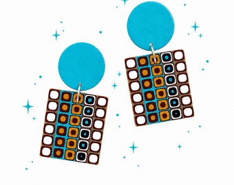 May include: A pair of retro-style earrings. Each earring features a turquoise blue circle connected to a rectangular pendant. The pendant has a brown border and a pattern of white, brown, orange, and blue squares. The earrings are suspended by silver hooks.