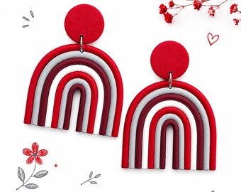 Puede incluir: Un par de pendientes en forma de arcoíris, rojos, blancos y burdeos. Cada pendiente presenta un círculo rojo y un diseño arqueado en capas. Sobre un fondo blanco con elementos decorativos.