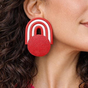 May include: A bold, red and white statement earring. The earring features a red textured circle beneath a red and white arched design. The earring is a unique, geometric accessory.