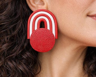 Puede incluir: Un llamativo pendiente rojo y blanco. El pendiente presenta un círculo rojo texturizado debajo de un diseño arqueado rojo y blanco. Un accesorio geométrico único.