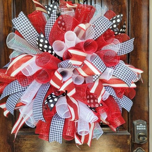 May include: A decorative wreath featuring red, white, and black ribbons in various patterns, including polka dots, houndstooth, and stripes. The wreath is made of mesh and ribbons, creating a full, textured appearance. It is displayed on a wooden door.