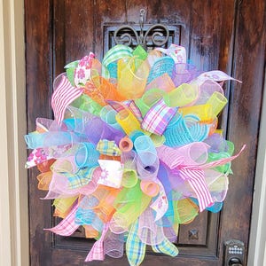 May include: A colorful wreath made of mesh and ribbon. The wreath is decorated with various colors, including pink, purple, blue, green, yellow, and orange. The wreath is hanging on a wooden door.